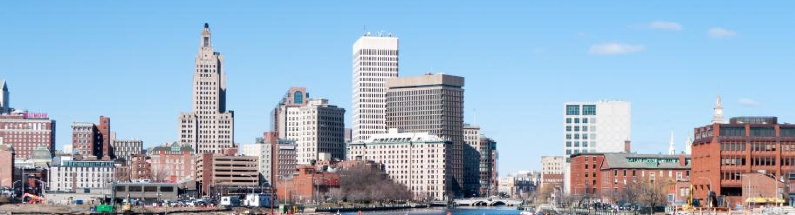 Providence Rhode Island skyline, water in foreground, large buildings in background, small clouds in blue sky above