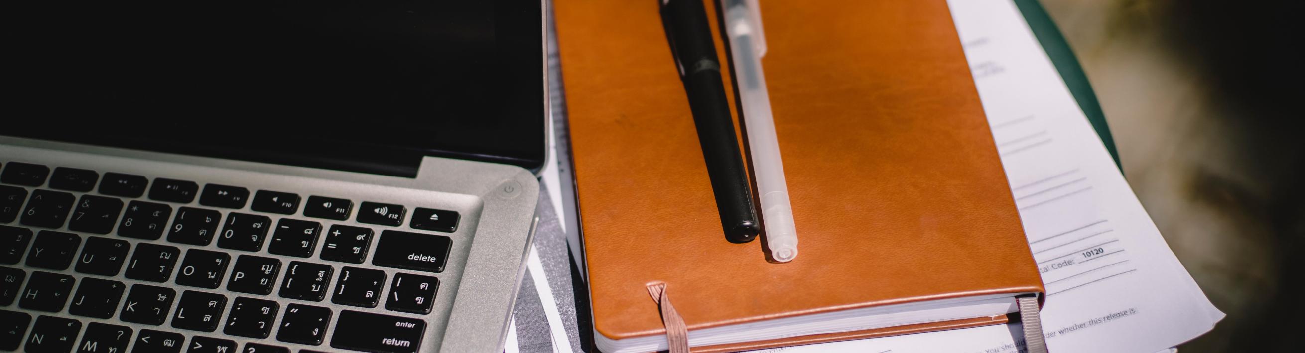 A stack of papers under a notepad and two pens next to a laptop.